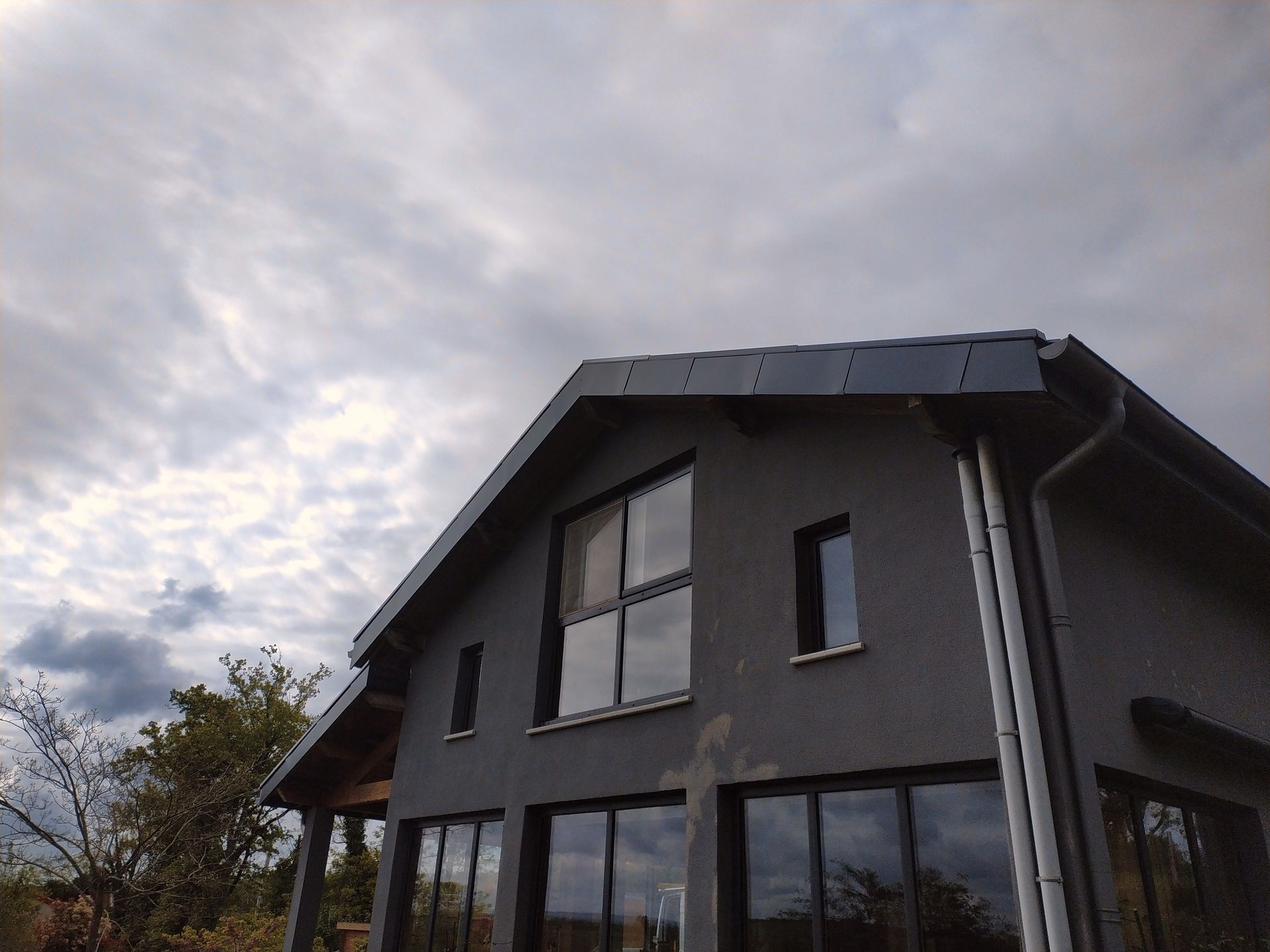 Maison à deux étages avec extérieur en stuc gris, grandes fenêtres et ciel nuageux.