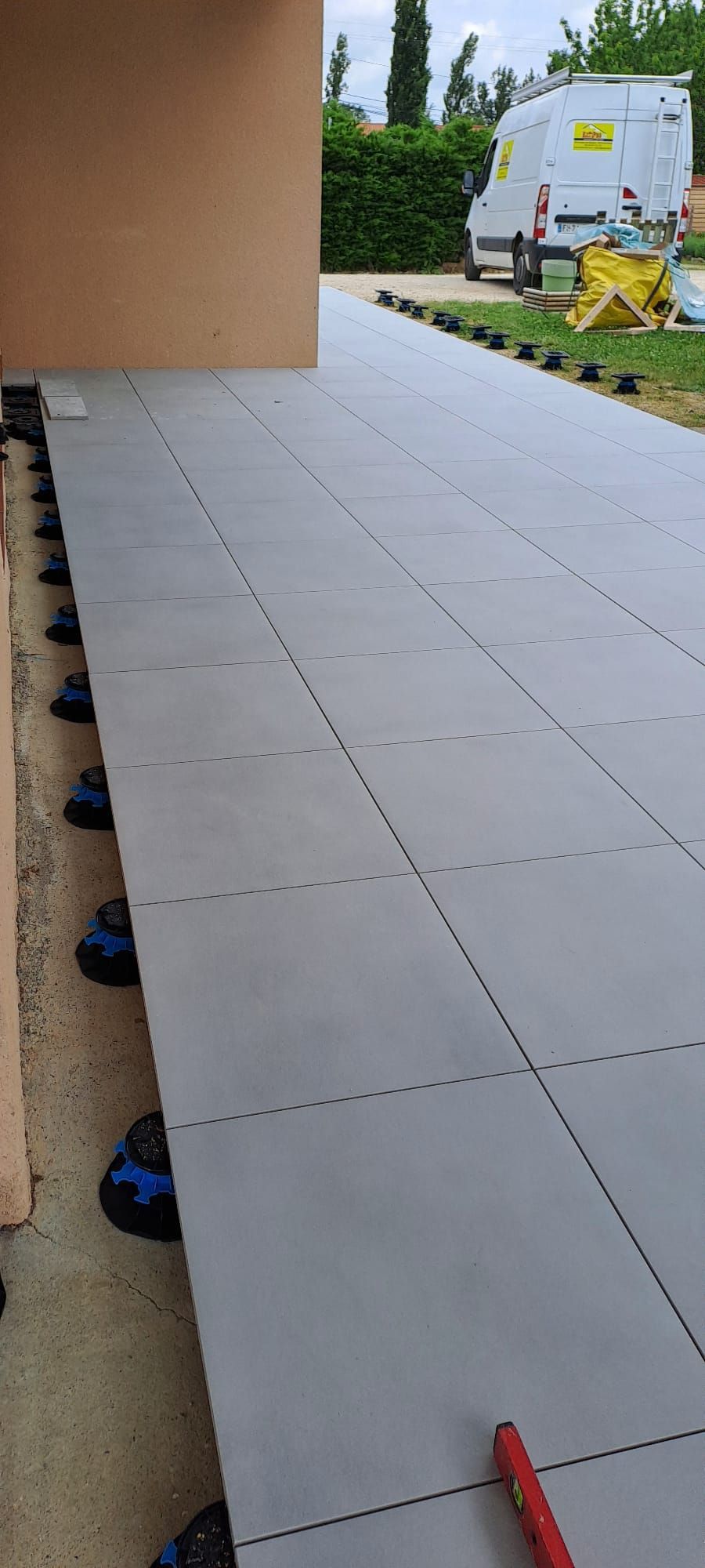 Un patio carrelé gris en cours de construction à côté d'un bâtiment marron clair avec une camionnette en arrière-plan.