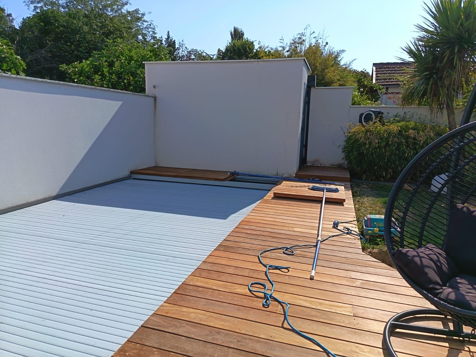 Terrasse en bois et patio pavé avec un mur, des arbres et une chaise suspendue dans une cour arrière par une journée ensoleillée.