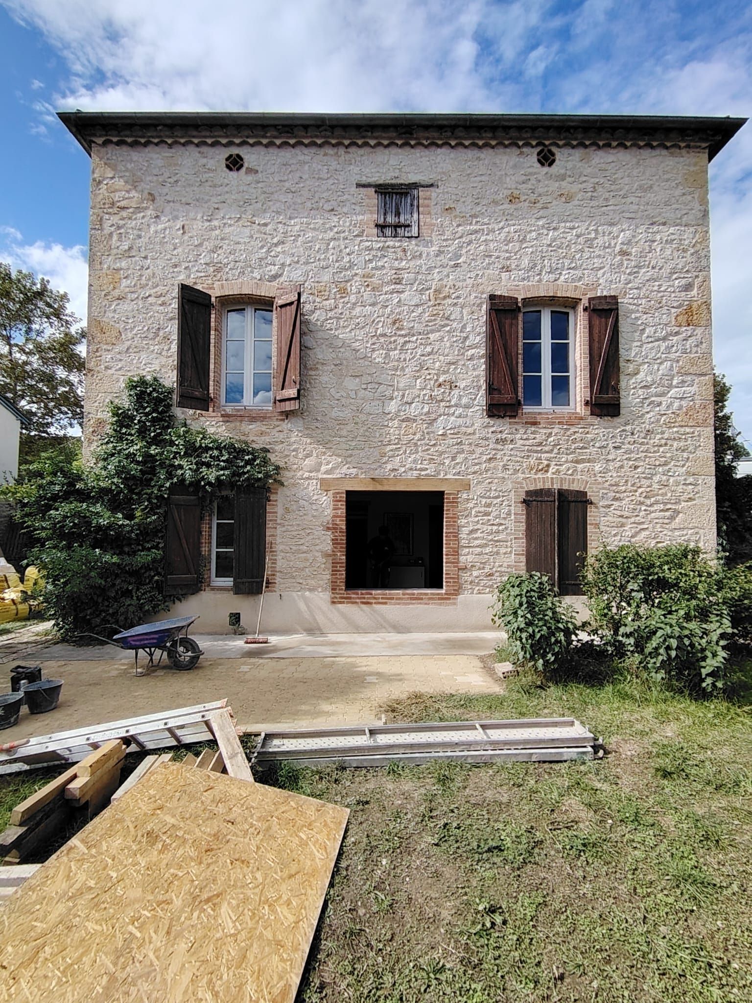 Maison en pierre avec porte ouverte, volets et matériaux de construction à l'extérieur par une journée ensoleillée.
