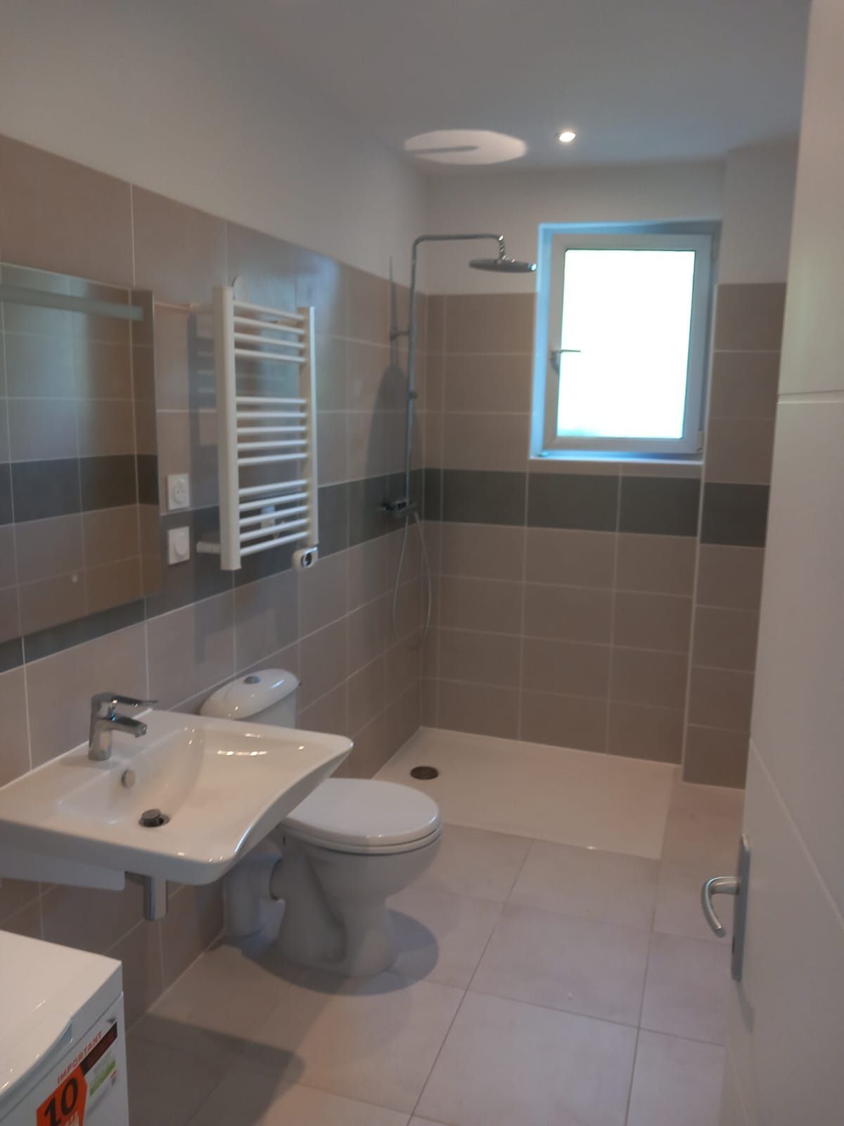 Salle de bain moderne avec toilettes, lavabo, douche et sèche-serviettes. Couleurs neutres et fenêtre.