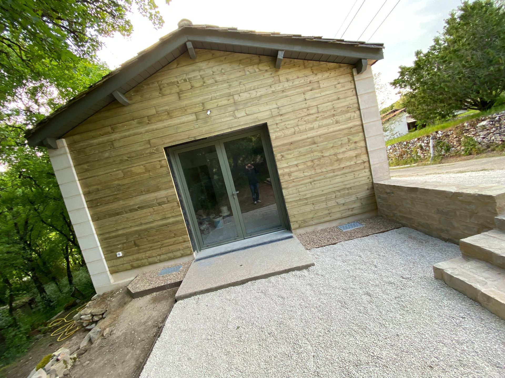 Bâtiment en bois avec portes vitrées et patio en gravier, niché près des arbres.