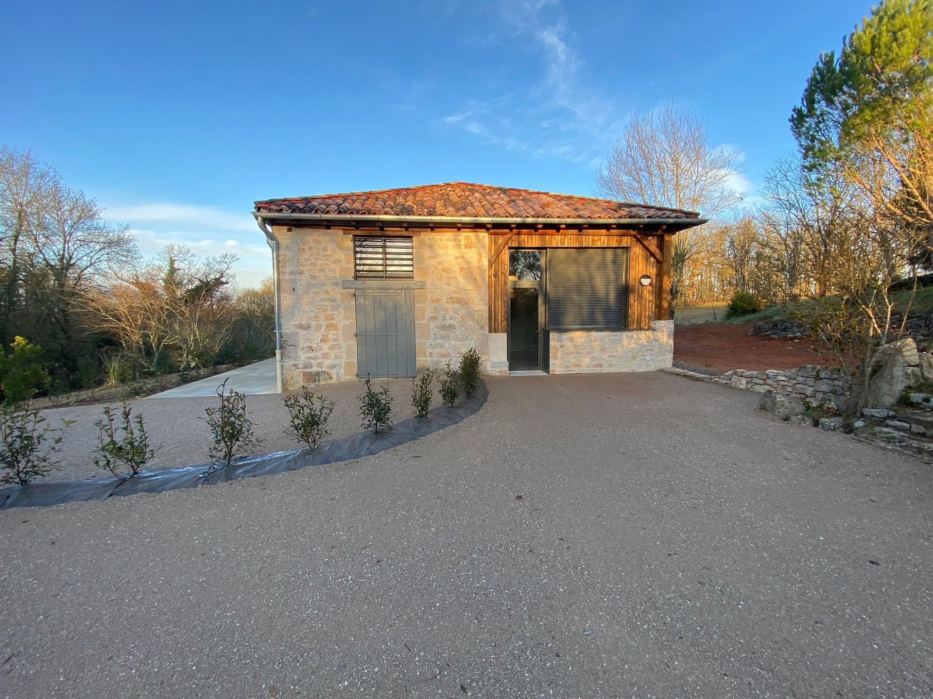 Chalet en pierre avec allée de gravier, arbres et ciel bleu.
