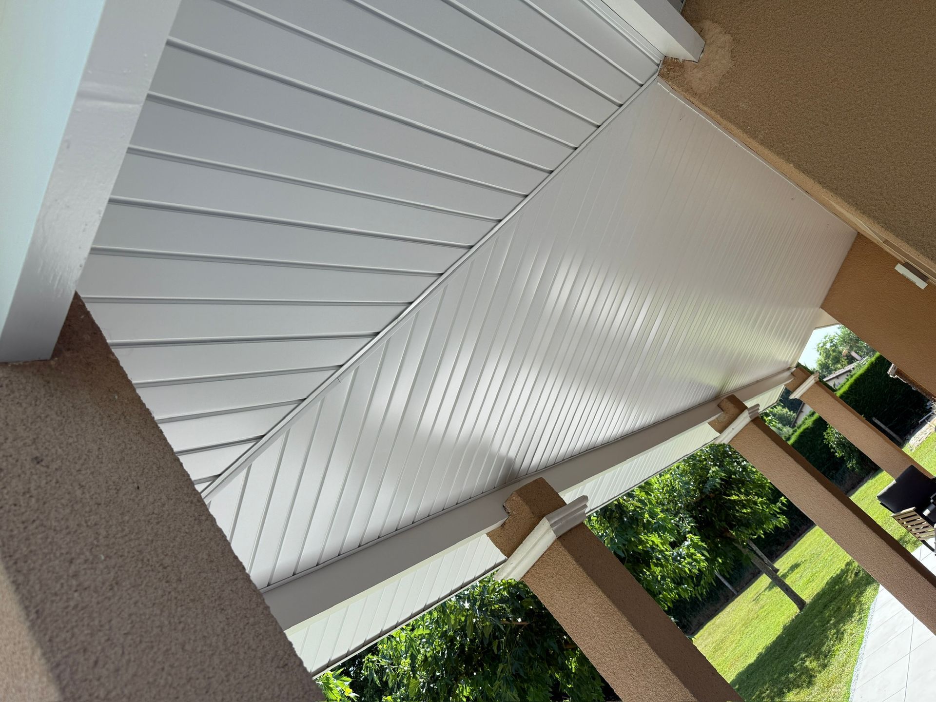 Plafond de patio à panneaux blancs avec colonnes beiges et arbres vert clair en arrière-plan.