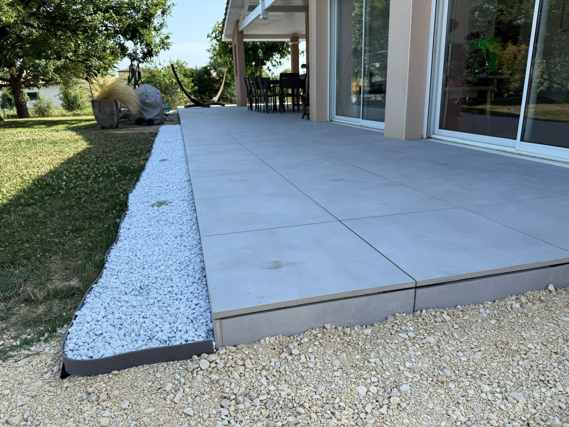 Patio gris avec bordure de gravier blanc, adjacent à la pelouse et à la maison avec portes vitrées.