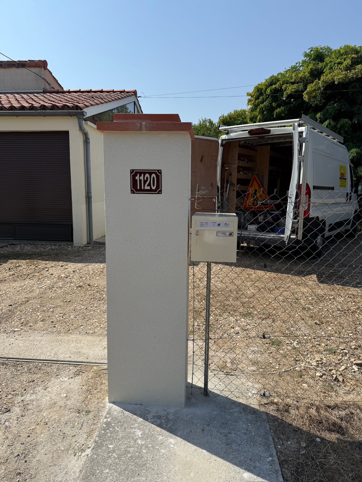 Une boîte aux lettres portant le numéro « 1120 » sur un pilier en béton. Une camionnette est garée derrière.