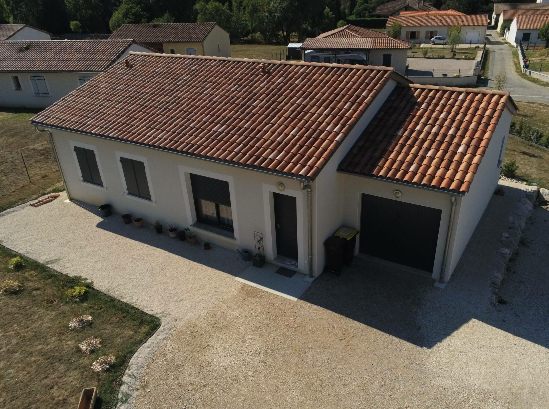 Maison blanche avec toit en tuiles rouges, allée en gravier et porte de garage dans un cadre suburbain ensoleillé.