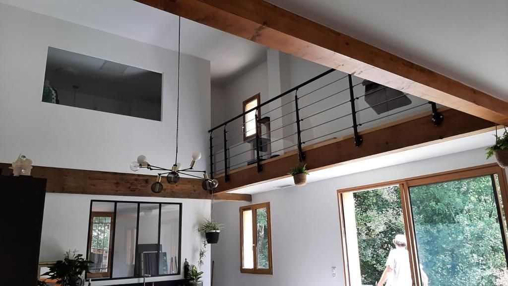 Intérieur avec mezzanine, poutres en bois, balustrade noire et grandes fenêtres donnant sur la verdure.