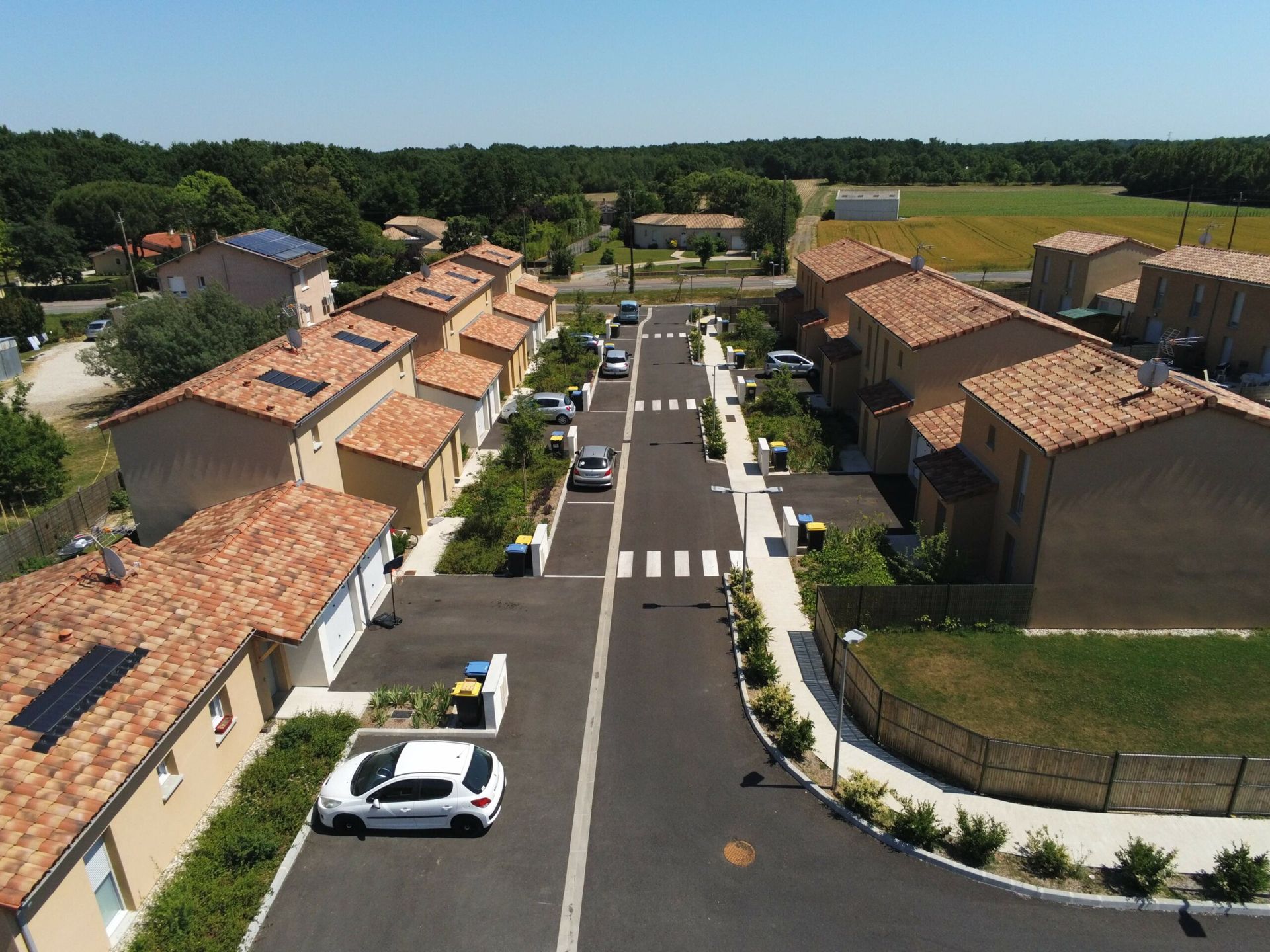 Vue aérienne d'une rue bordée de maisons, de toits de tuiles rouges et de voitures garées. Champs verdoyants et arbres en arrière-plan.