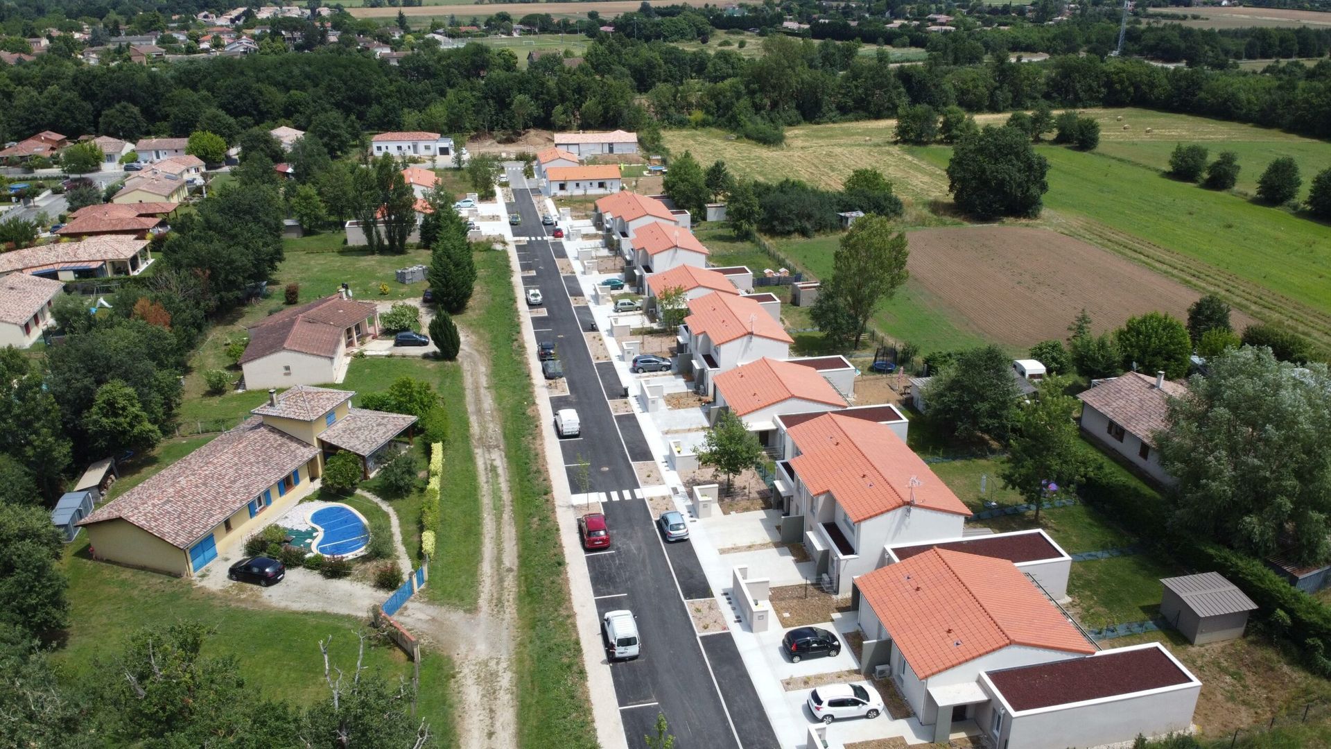 Vue aérienne d'une rue résidentielle avec des maisons nouvellement construites, des pelouses vertes et des champs au-delà, avec des voitures sur la route.