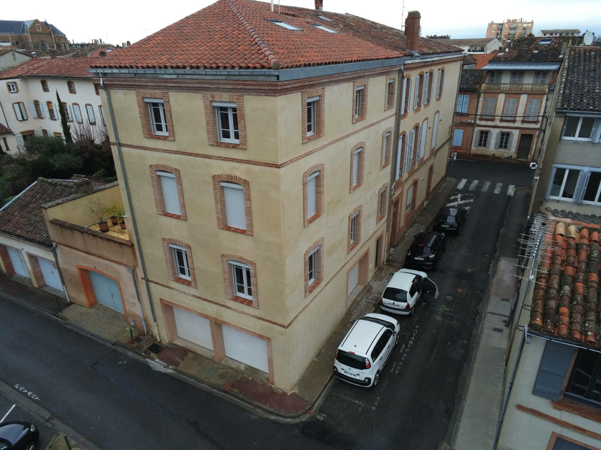 Bâtiment d'angle avec murs beiges, toit en tuiles rouges et voitures garées dans la rue.