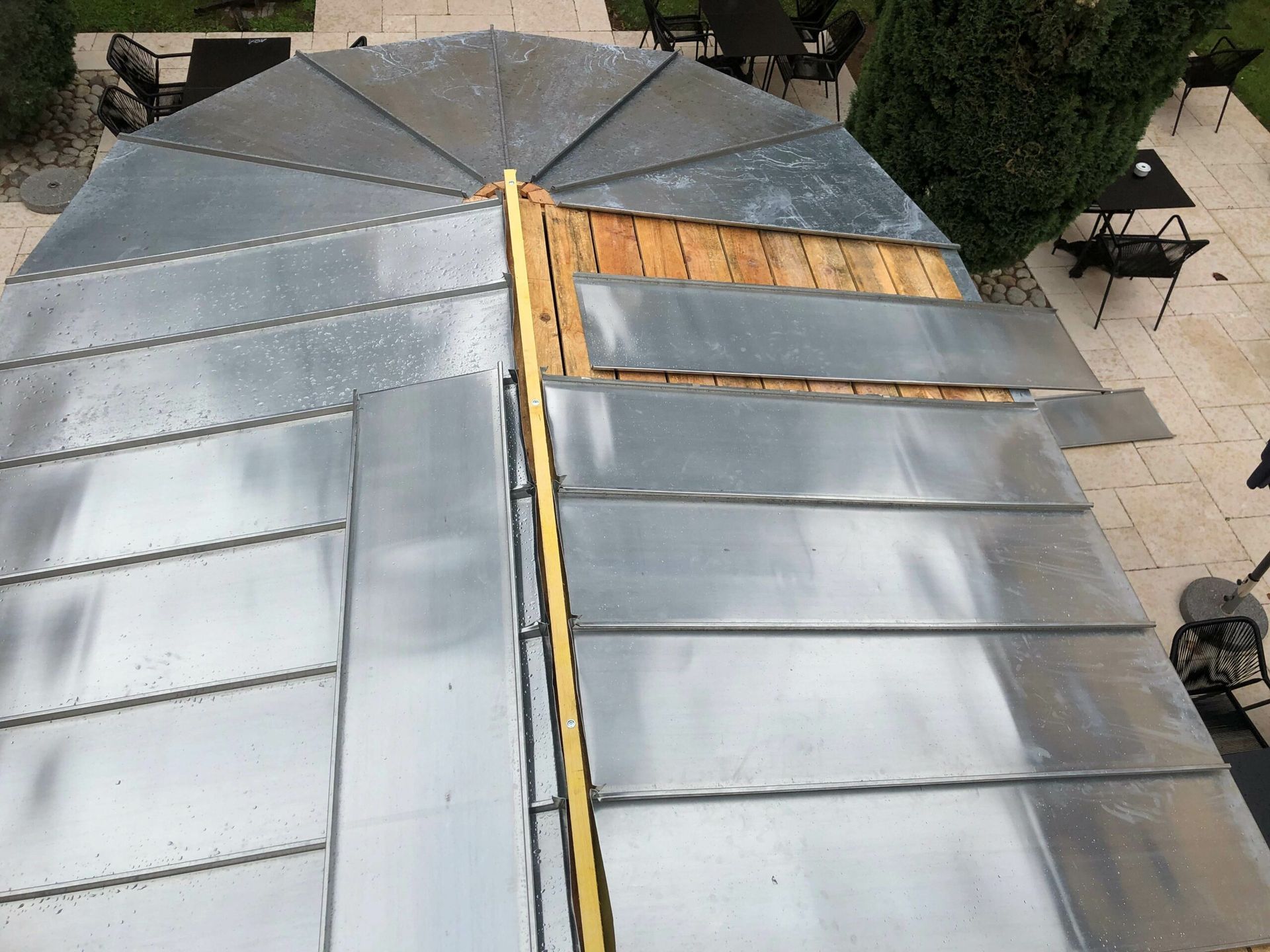 Toiture métallique partiellement installée sur un gazébo ; métal argenté, poutres de bois, aménagement extérieur.