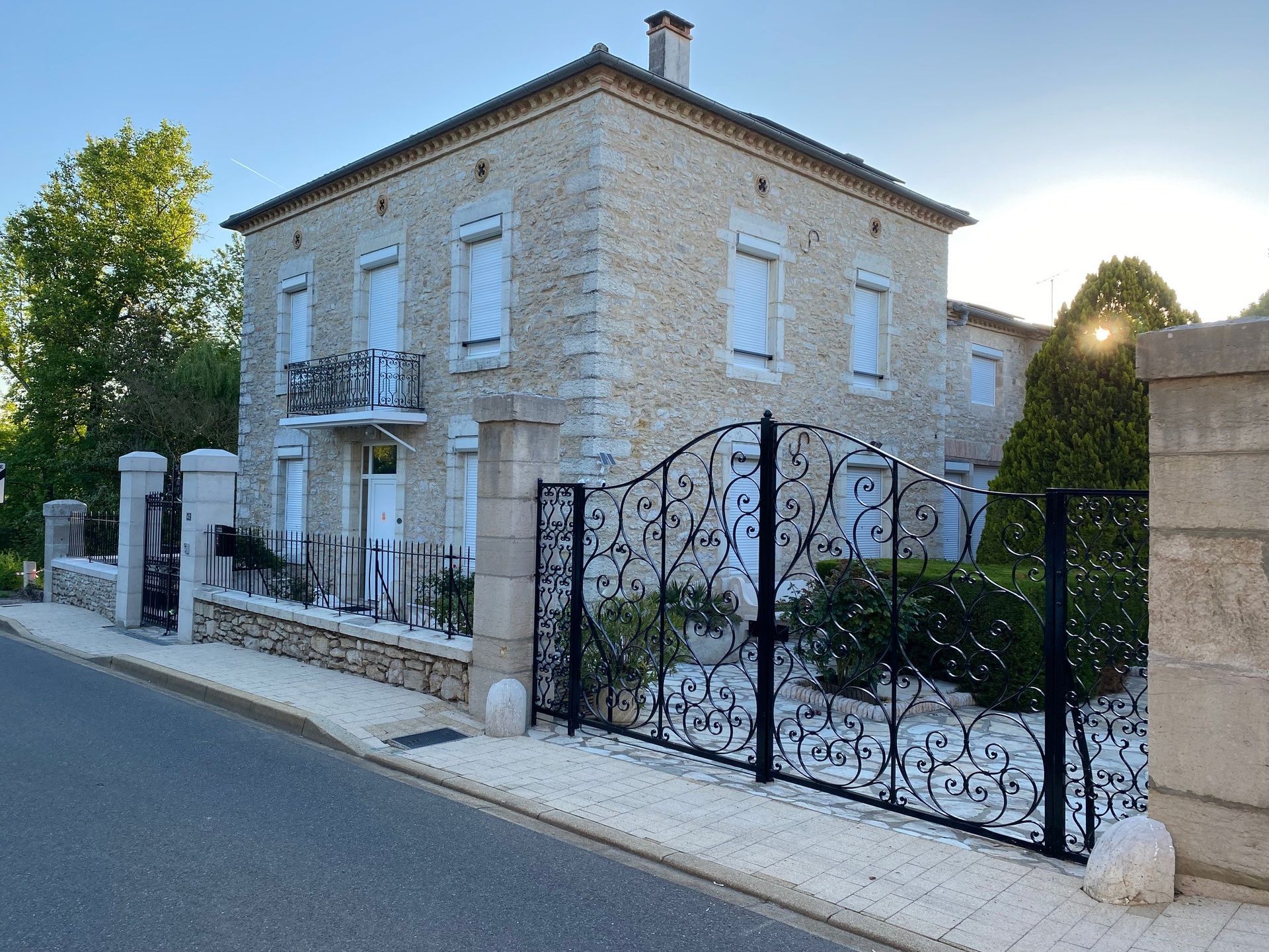 Maison en pierre avec portail en fer noir, volets blancs et balcon, dans une rue.