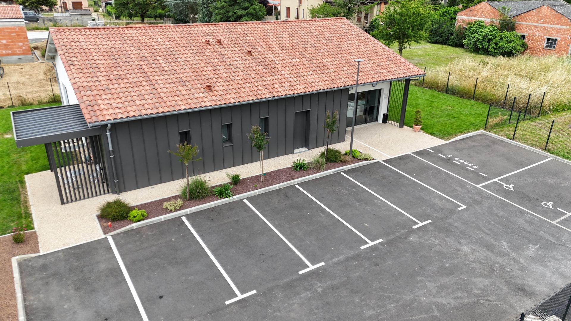 Bâtiment avec revêtement gris foncé, toiture en terre cuite et stationnement.