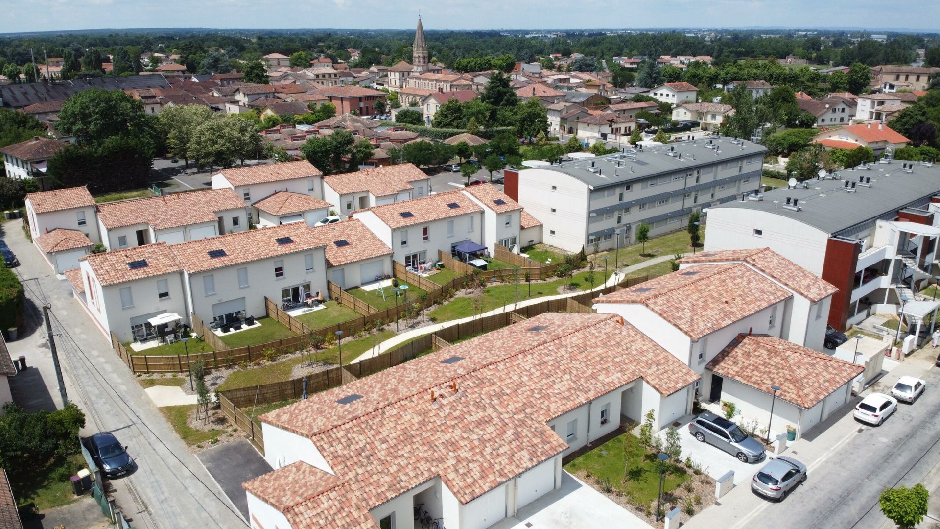 Vue aérienne d'un quartier résidentiel avec des maisons blanches, des toits de tuiles rouges et un clocher d'église au loin.
