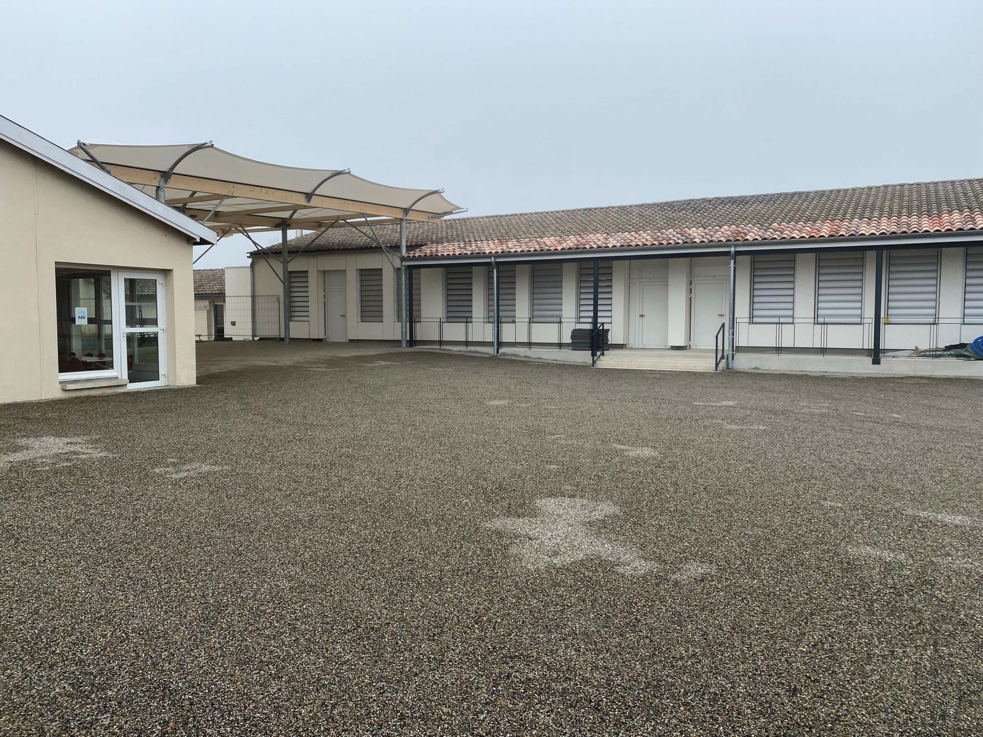 Vue extérieure de bâtiments aux murs blancs, au sol en gravier et sous un ciel couvert.