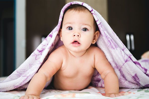 Un bébé qui sort la tête de sous une couverture violette sur un lit, l'air surpris.