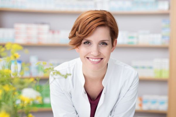 Pharmacien en blouse blanche souriant à la caméra ; étagères de pharmacie en arrière-plan.