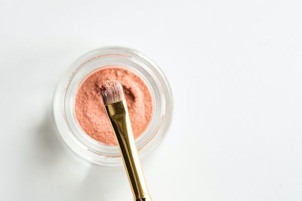 Vue de dessus d'un pot de fard à paupières couleur pêche avec un pinceau de maquillage trempé dedans, sur fond blanc.