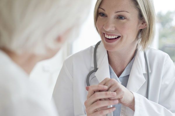 Une doctoresse blonde sourit en parlant avec un patient âgé. Elle porte une blouse blanche et un stéthoscope. À l'intérieur.