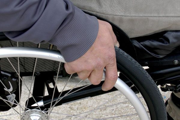 Gros plan sur la main d'une personne posée sur une roue de fauteuil roulant. Veste grise, pantalon beige. Mise au point sur la main.