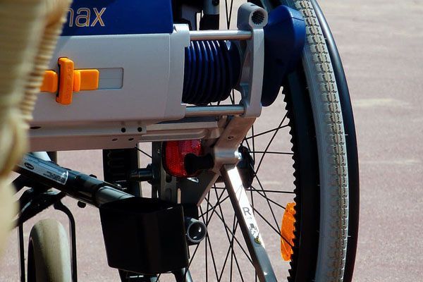 Gros plan sur le moteur et la roue d'un fauteuil roulant, avec des composants bleus, blancs, noirs et orange, en extérieur.