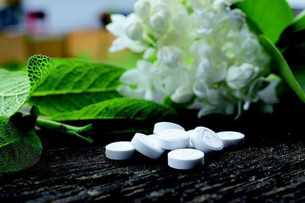 Des pilules blanches éparpillées sur du bois sombre, avec des feuilles et des fleurs blanches en arrière-plan.
