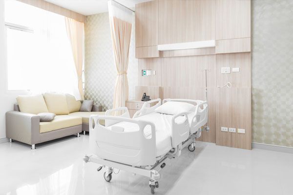 Chambre d'hôpital avec un lit, un canapé et des boiseries claires ; fenêtre avec des rideaux transparents.