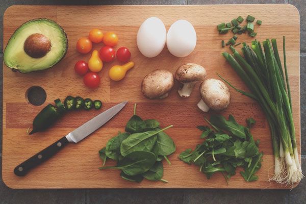 Planche à découper en bois avec avocat, tomates, œufs, champignons, légumes verts et un couteau.