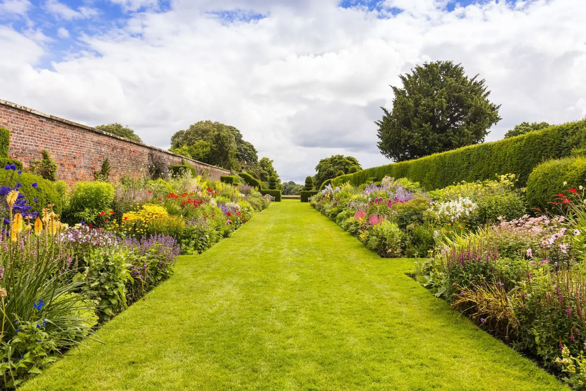 Allée de jardin soignée bordée de fleurs colorées et de haies sous un ciel nuageux.