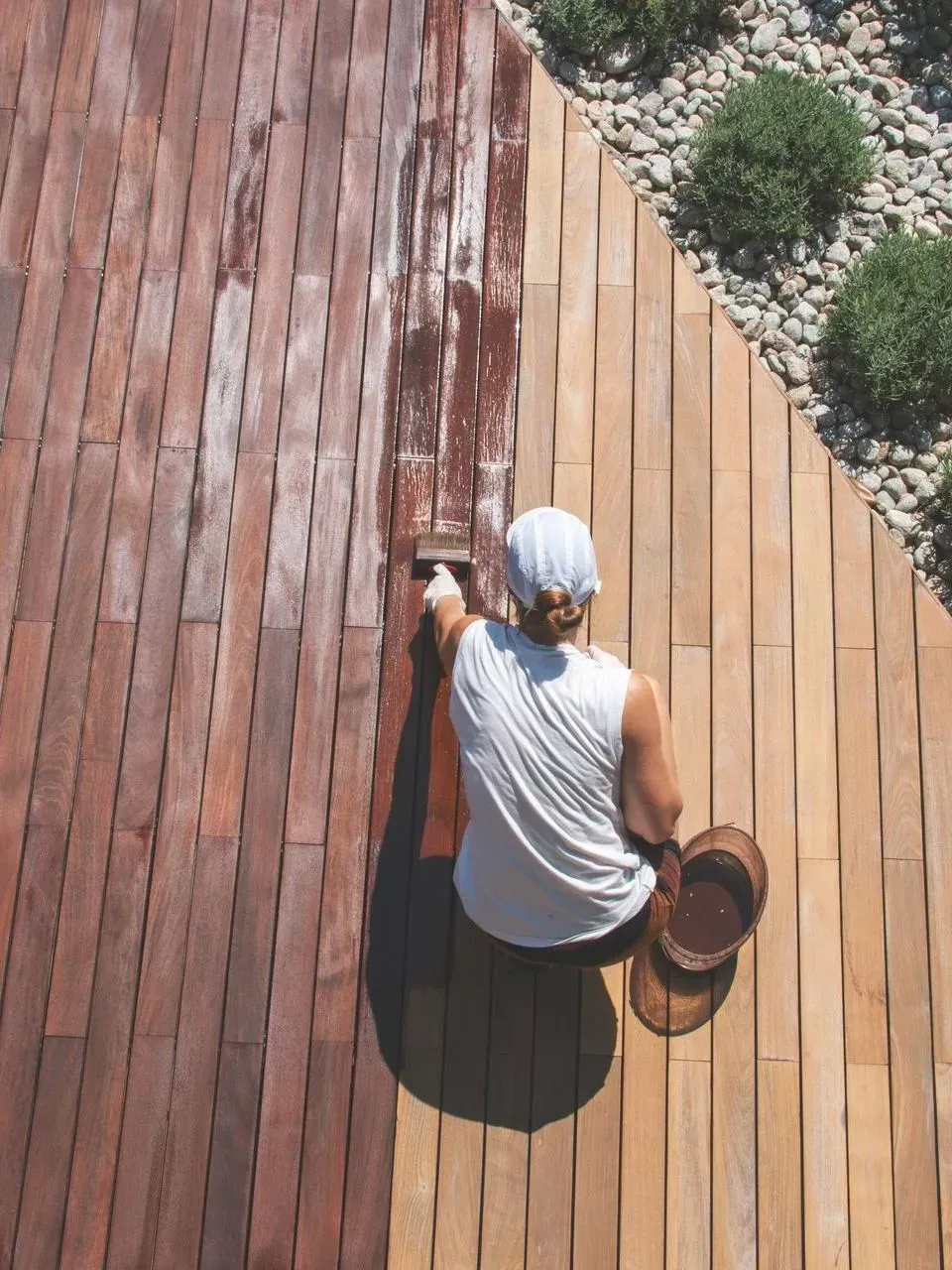 Une personne peint une terrasse en bois patinée par le temps, bordée de gravier et d'arbustes, sous un soleil éclatant.