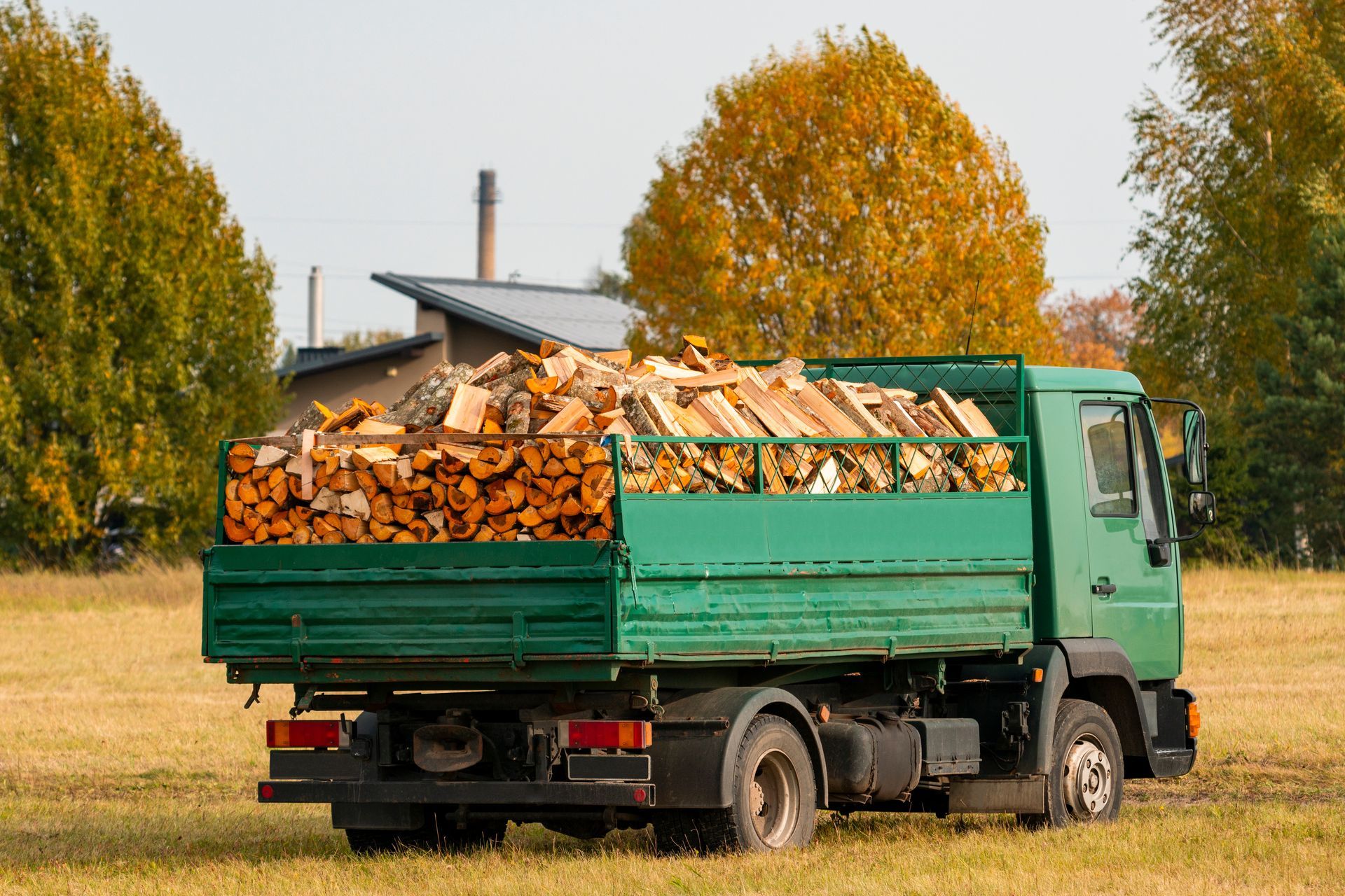 Un camion vert chargé de bois de chauffage dans un champ, avec des arbres d'automne en arrière-plan.