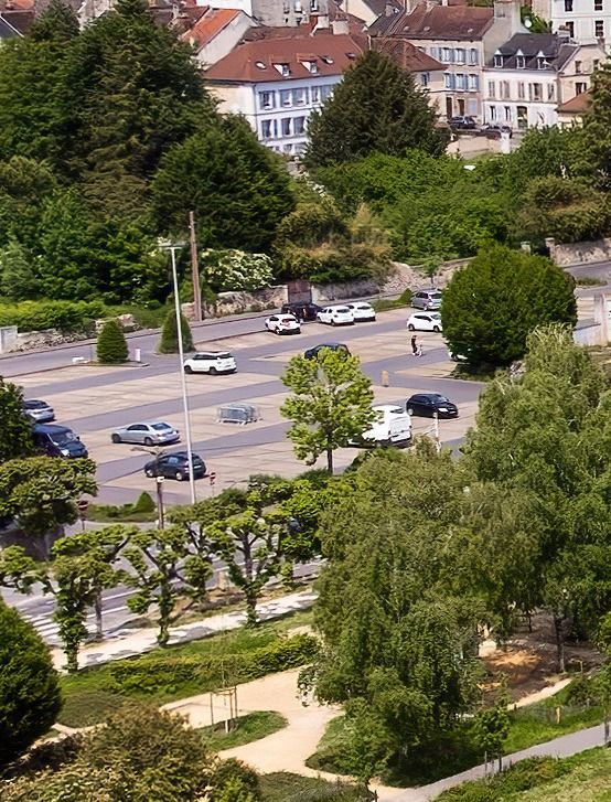 Parking bordé d'arbres avec plusieurs voitures, vu du ciel, avec des bâtiments en arrière-plan.