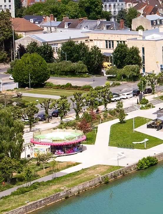 Parc riverain avec des sentiers sinueux, des arbres et un parterre de fleurs roses bordant une promenade au bord de l'eau.