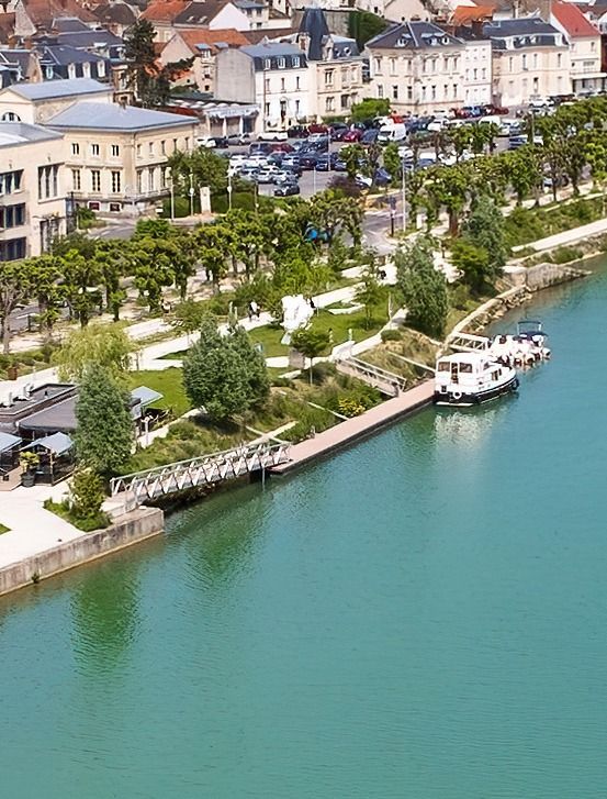 Une promenade au bord de la rivière avec des arbres, des bâtiments blancs et des bateaux sur une eau turquoise dans une ville côtière.