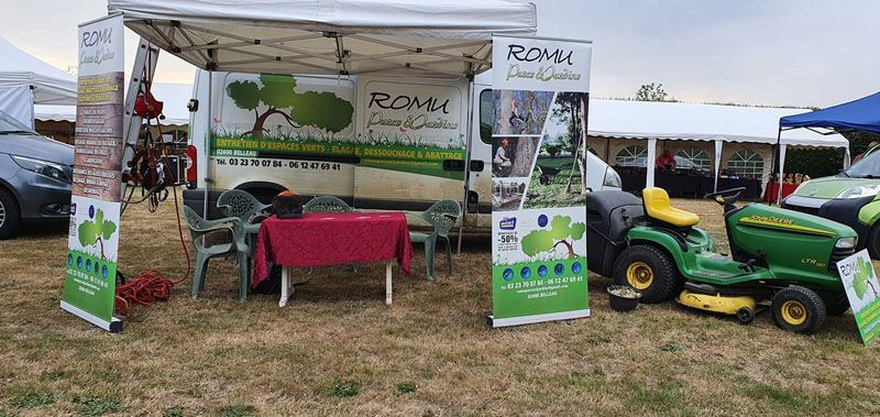 Stand promotionnel extérieur avec exposition de tracteurs verts et banderole sous un auvent blanc sur un terrain herbeux.