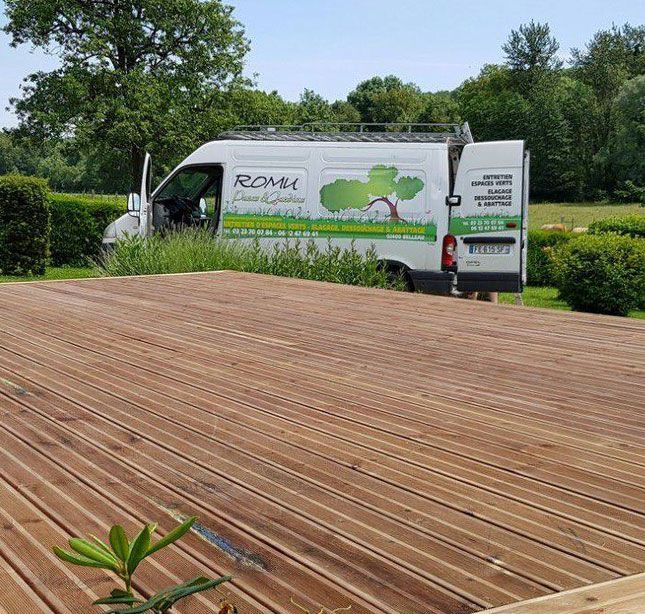 Une camionnette de livraison blanche avec un logo vert est garée à côté d'une terrasse en bois dans un parc gazonné.