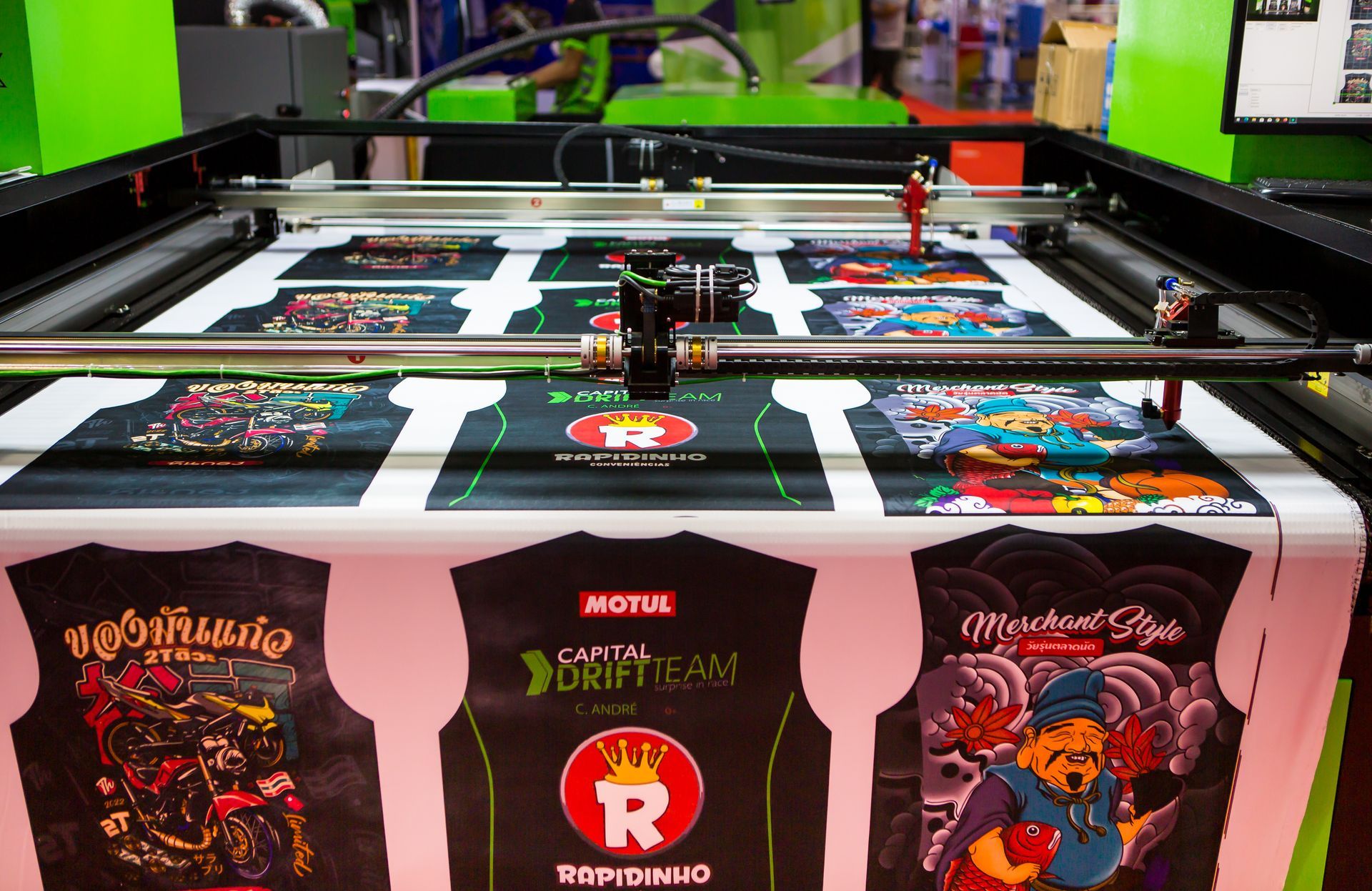 Une machine d'impression découpe des motifs sur du tissu. Plusieurs modèles de gilets de moto imprimés sont exposés.