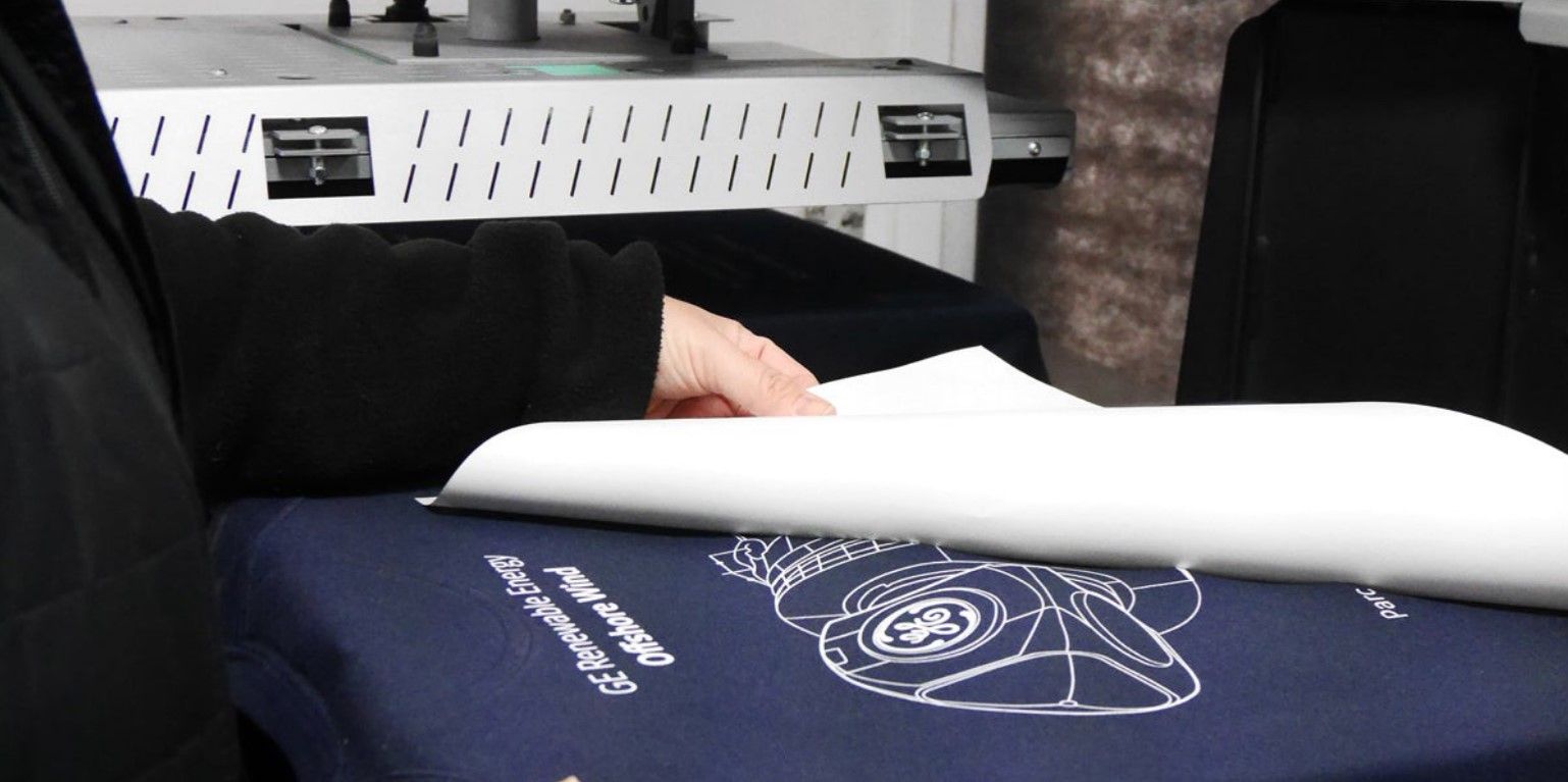 Une personne pose du papier de transfert sérigraphique sur un tshirt bleu, près d'une machine à imprimer.
