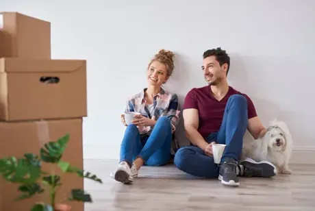 Una pareja y un perro pequeño sentados contra una pared cerca de unas cajas, con aspecto satisfecho. Una mujer sostiene una taza, un hombre sonríe, en el interior.