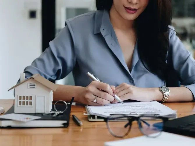 Una mujer con una camisa azul firma documentos en un escritorio con una maqueta de casa, gafas y llaves.