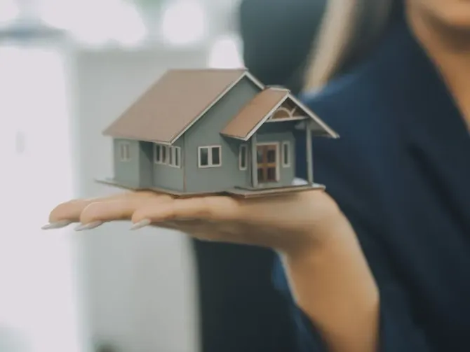 Mujer sosteniendo una casa en miniatura, posiblemente una presentación inmobiliaria. La casa es gris con techo marrón; la mujer lleva un blazer oscuro.