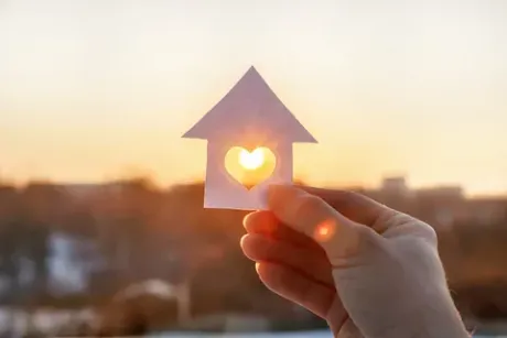 Mano sosteniendo una casa de papel blanco con una ventana en forma de corazón contra un sol poniente.