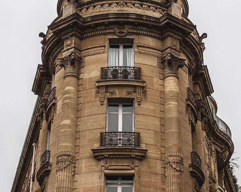 Angle d'un bâtiment en pierre beige aux balcons ornés et au toit arrondi, se détachant sur un ciel nuageux.