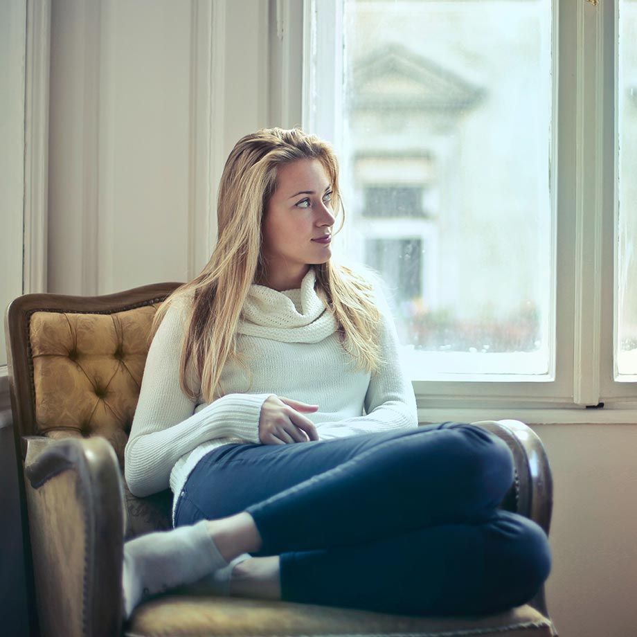 Femme en pull blanc et pantalon bleu assise sur une chaise, regardant par la fenêtre.