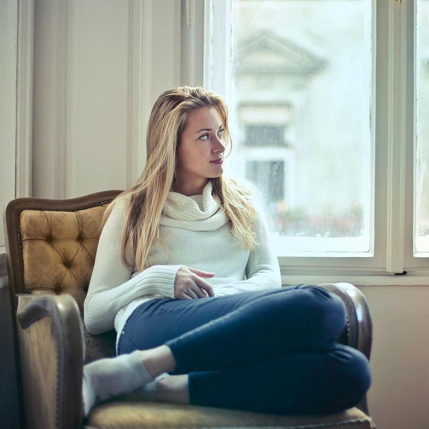 Femme en pull blanc et pantalon bleu assise sur une chaise, regardant par la fenêtre.