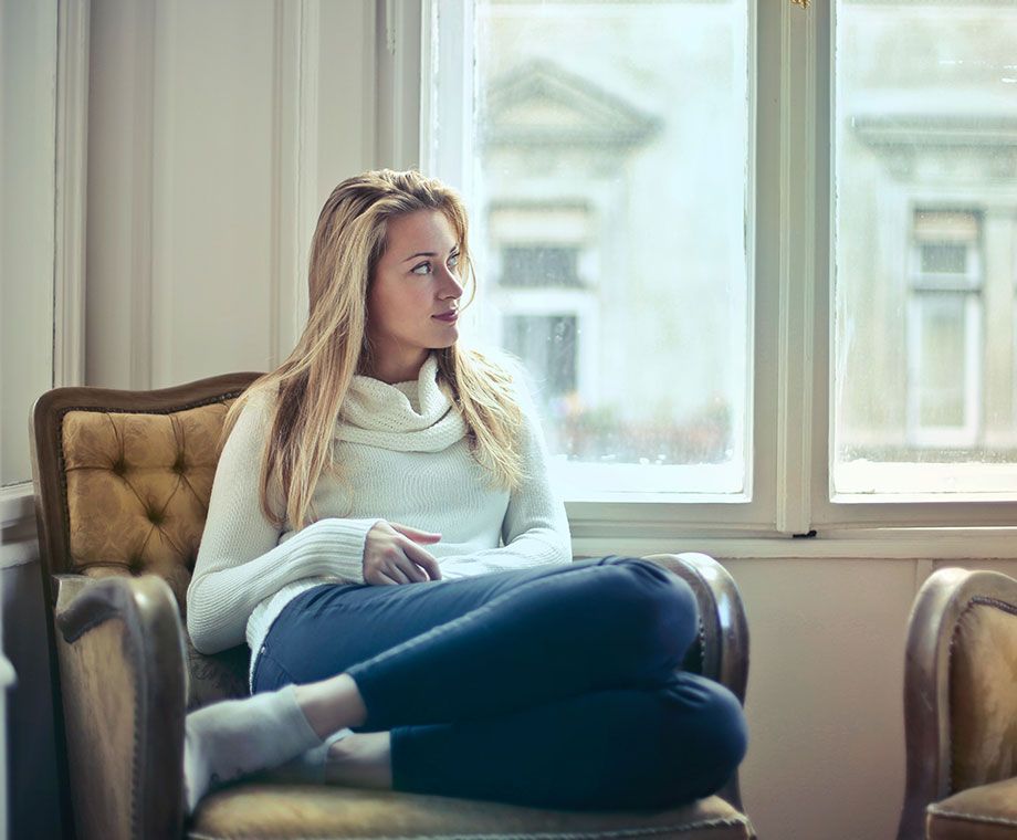 Une femme en pull blanc et pantalon bleu est assise sur une chaise et regarde par la fenêtre.