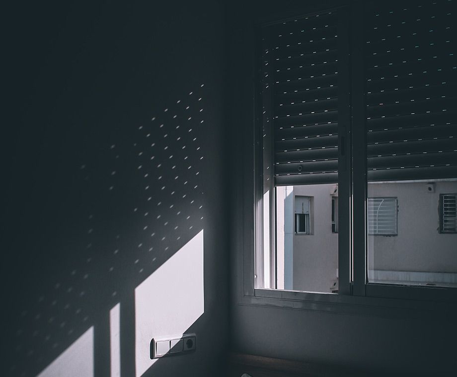 La lumière du soleil filtrée par les stores fermés projette des motifs pointillés sur un mur blanc d'une pièce.
