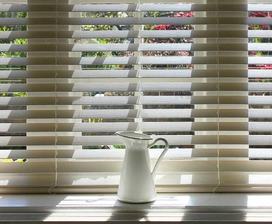 Pichet blanc sur le rebord de la fenêtre, lumière du soleil filtrant à travers les stores ouverts, feuillage vert et rouge flou visible à l'extérieur.