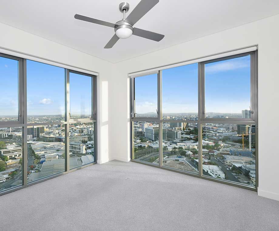 Chambre vide avec baies vitrées offrant une vue sur la ville, moquette claire, ventilateur de plafond.
